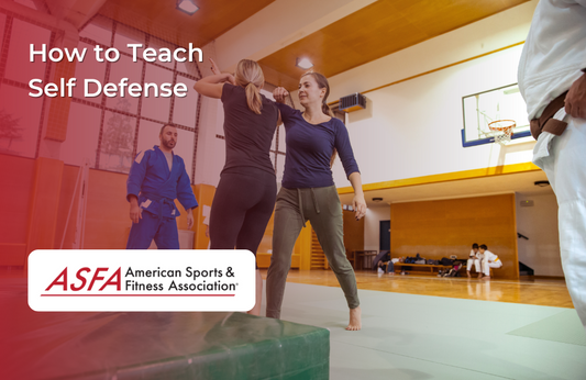 Two women practice a self-defense technique in a gym while others observe, representing a self-defense training session.