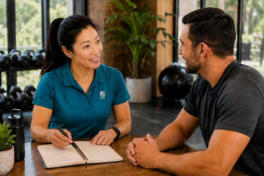 ASFA advanced health & wellness coach in a teal polo shirt with an ASFA logo sits at a wooden table, writing in a notebook while talking to a male client in a gym setting.