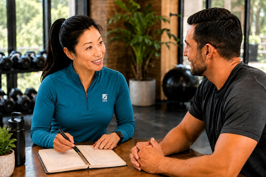 ASFA advanced health & wellness coach in a teal polo shirt with an ASFA logo sits at a wooden table, writing in a notebook while talking to a male client in a gym setting.