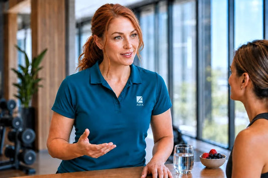 ASFA wellness coach in a teal polo shirt with a visible  logo is talking to another woman at a table in a bright modern indoor setting with large windows.