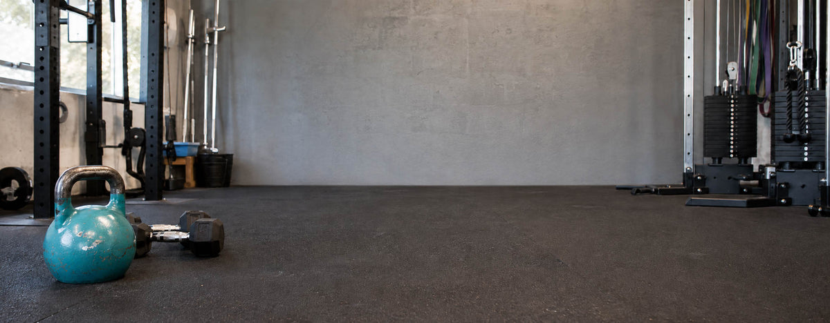 A teal kettlebell and a small dumbbell resting on dark gym flooring, with exercise equipment and a concrete wall in the background of an indoor gym space.
