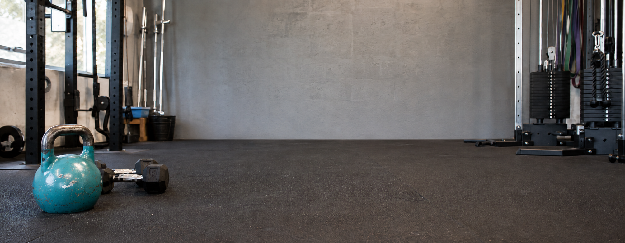 A teal kettlebell and a small dumbbell resting on dark gym flooring, with exercise equipment and a concrete wall in the background of an indoor gym space.