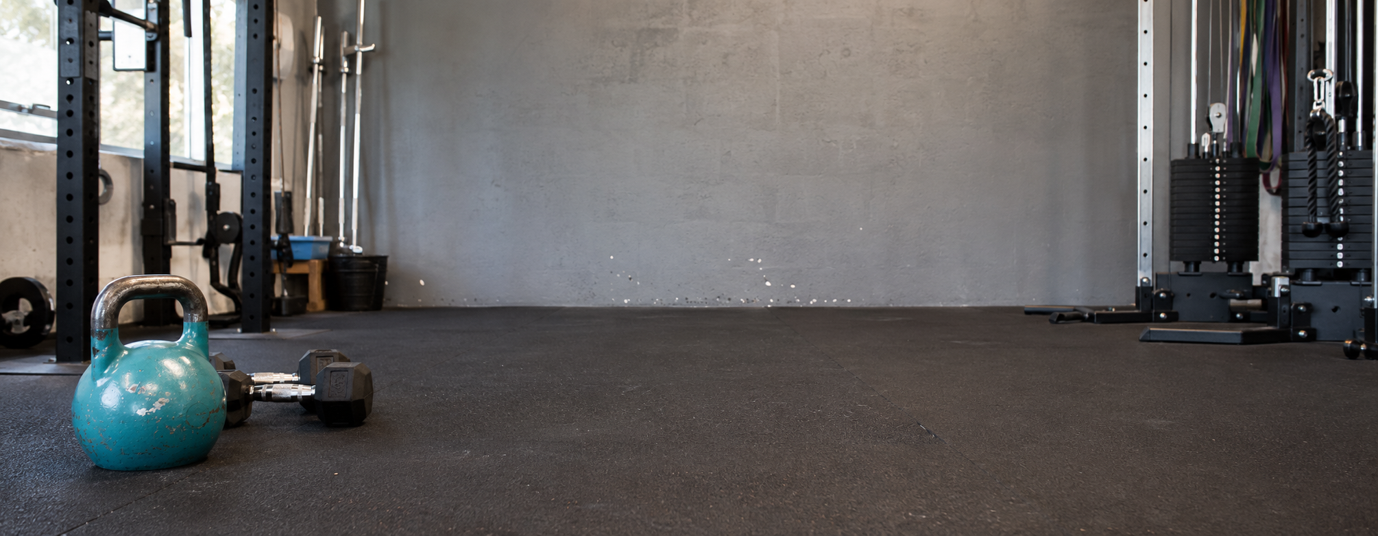 A teal kettlebell and a small dumbbell resting on dark gym flooring, with exercise equipment and a concrete wall in the background of an indoor gym space.